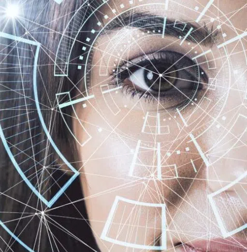 The image presents a close-up of part of a human face, focusing on one eye, overlaid with futuristic digital interface graphics. The eye is sharply in focus, while the rest of the face is partially cropped and softly blurred, drawing attention to the iris and surrounding area. Across the image, translucent geometric lines and nodes form a network-like pattern, suggesting data connections or artificial intelligence systems. Circular interface elements and targeting-style graphics are layered around the eye, resembling biometric scanning or facial recognition technology. The background transitions into a gradient of dark blue and grey tones, reinforcing a high-tech atmosphere. The combination of the human feature and digital overlays creates a visual theme centered on advanced technology, data analysis, and biometric identification systems.