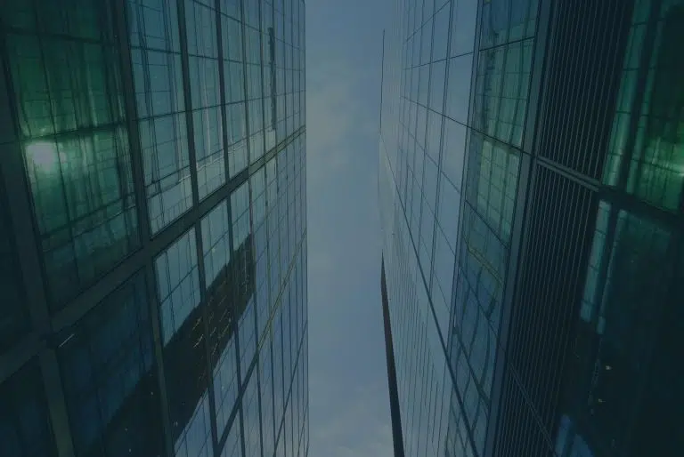 View looking upward between two tall, modern glass office buildings that rise on the left and right sides of the frame. The buildings have reflective glass façades with a grid of window panels, tinted blue-green, and they mirror parts of the surrounding city. The perspective creates strong vertical lines that converge toward a narrow strip of pale sky in the center. The overall scene feels symmetrical and geometric, with clean lines, cool tones, and a slightly overcast sky visible above.