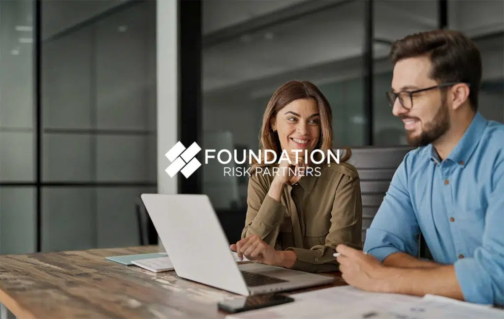 Two colleagues standing in a modern office discuss something on a tablet. A woman wearing a beige hijab and light blazer looks at the screen while a man in a light blue shirt holds the tablet and speaks. The overlay reads “Foundation Risk Partners” with the company logo.