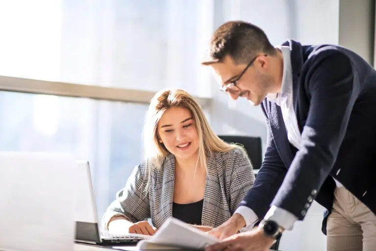 Two people in a bright office collaborate at a desk, reviewing documents while one points and explains.