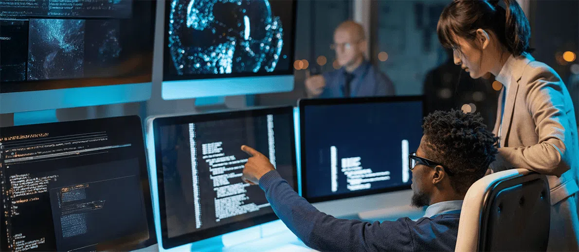 A team of cybersecurity or IT professionals works together in a dimly lit operations center, surrounded by large monitors displaying code, system data, and digital visualizations. One person seated at a workstation points toward a screen filled with lines of code while another stands beside them, observing and discussing the information.
