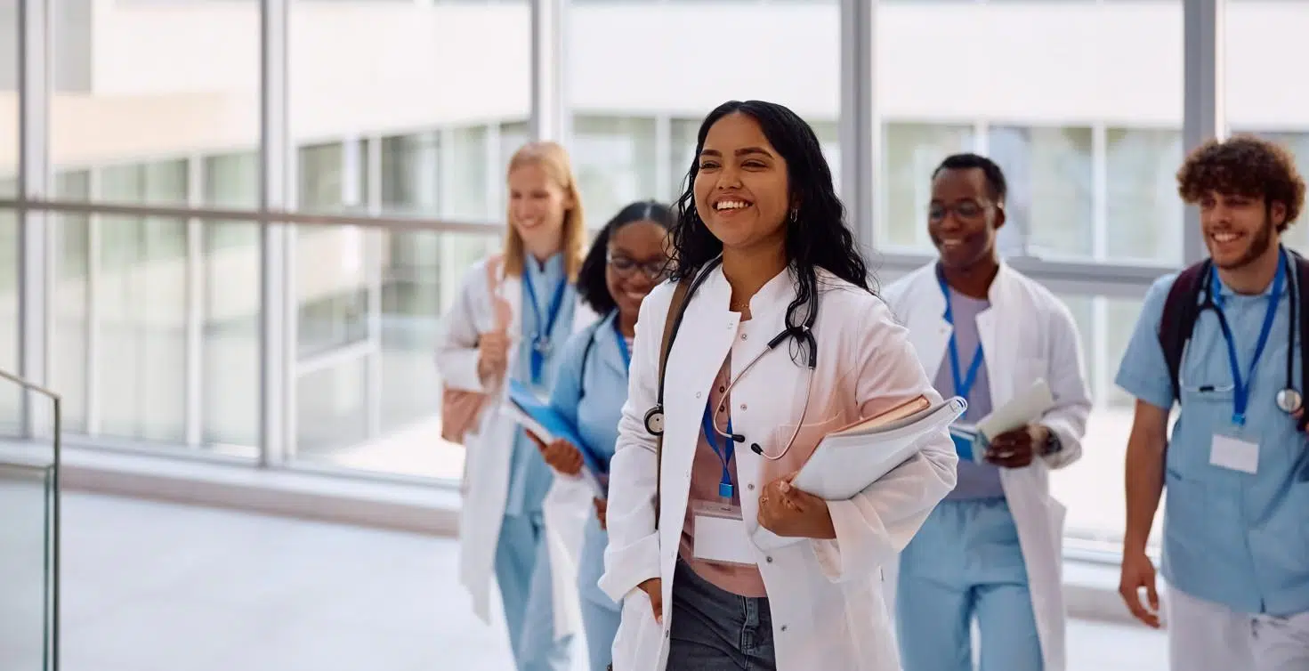 A group of healthcare professionals walking together down a bright, modern corridor with large windows and natural light. In the foreground, one individual wears a white medical coat and has a stethoscope around the neck, holding a folder or clipboard and wearing an ID badge on a lanyard. Behind, several others walk in the same direction, also wearing medical attire such as scrubs or white coats, many with stethoscopes and ID badges visible. Some carry notebooks or documents. The corridor appears clean and spacious, with glass panels and a view of another building outside. The overall scene shows a collaborative healthcare environment with multiple professionals moving together in a clinical setting.