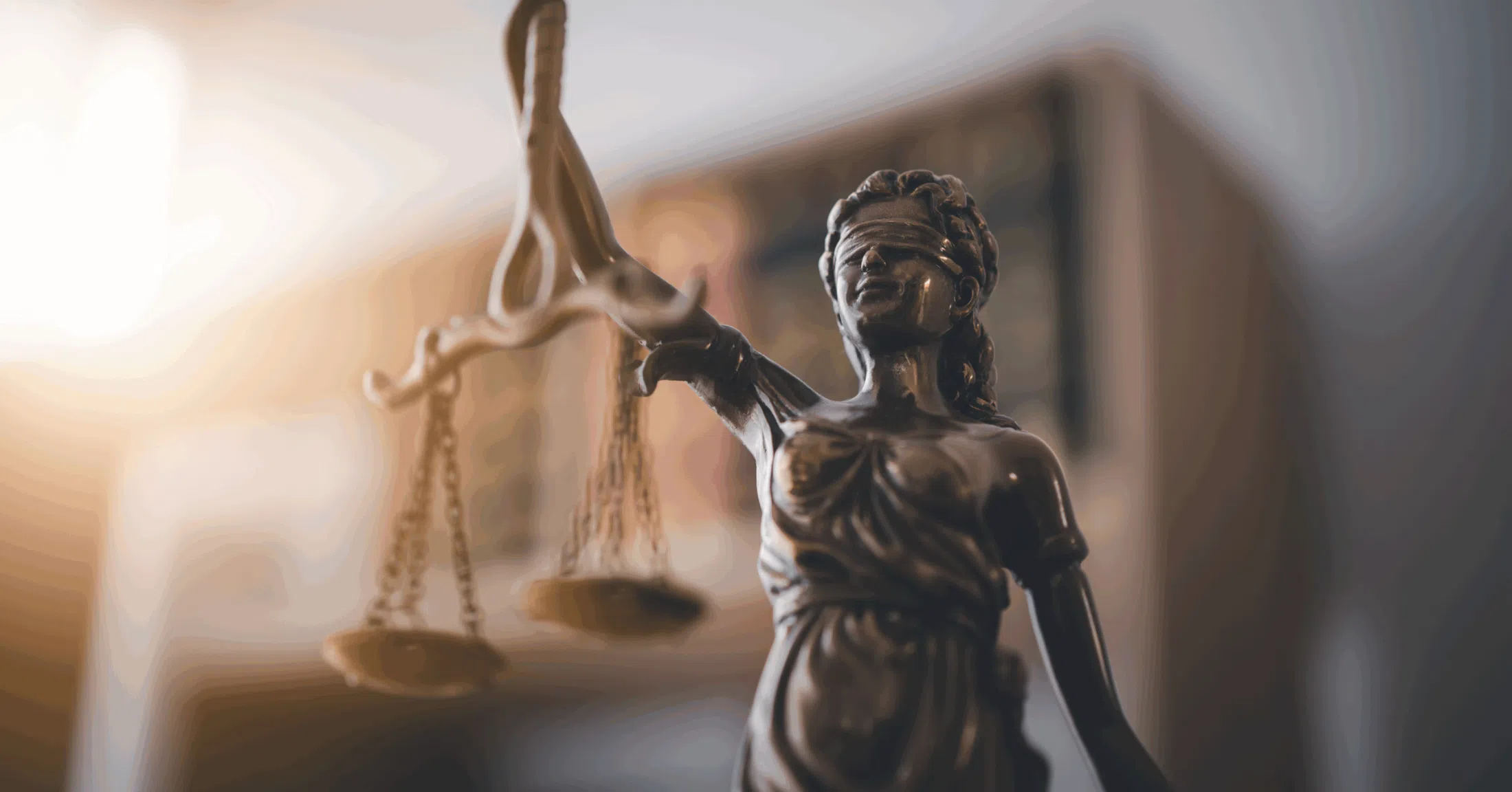 A close-up of a bronze statue representing Lady Justice standing indoors. The statue holds a set of balance scales suspended from one raised hand, with the chains and plates visible in the foreground. The figure wears a blindfold and flowing robes, and the metal surface has a dark, polished finish that reflects the warm ambient light. The background is softly blurred, showing indistinct shelves or furniture, creating a shallow depth-of-field effect that keeps the focus on the statue and its scales.