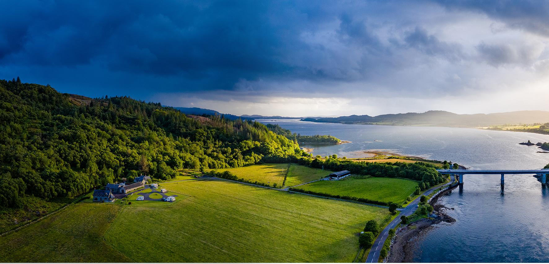 A wide, panoramic landscape view showing a coastal countryside with rolling green fields, dense woodland, and a large body of water stretching into the distance. In the foreground, a rural property with several buildings sits within open farmland bordered by hedgerows and narrow roads. To the right, a long bridge extends across the water, connecting land on either side. The shoreline curves gently around the bay, with patches of trees and small inlets visible. In the background, low hills and mountains fade into the horizon. The sky is dramatic, with dark storm clouds on the left gradually giving way to brighter light on the right, casting a mix of shadow and sunlight across the scene.