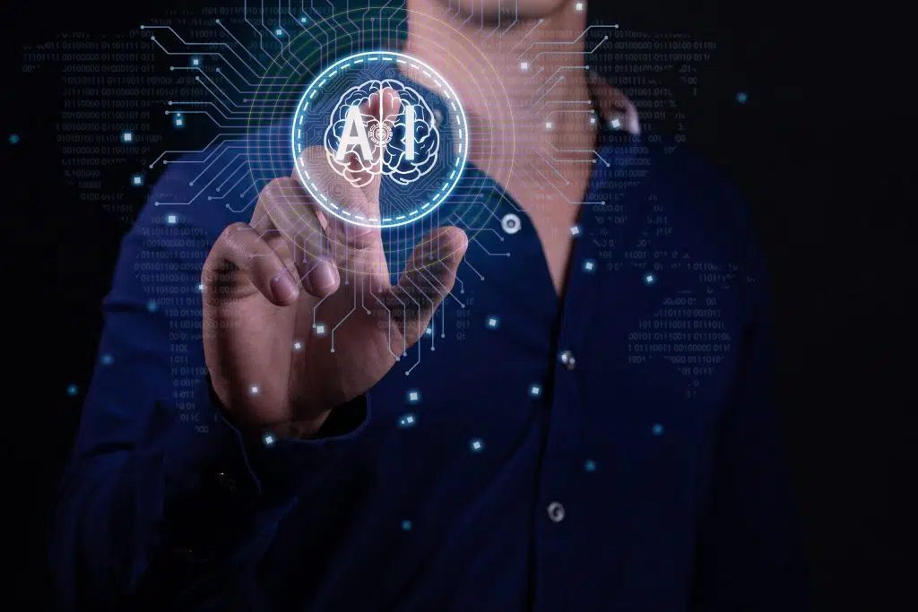 A person in a dark blue shirt is interacting with a futuristic holographic interface. They are pressing a virtual button that displays an icon of a brain within a circle, with the word AI in it. The background features digital binary code and circuit lines, suggesting advanced technology or computing concepts. There are glowing dots connected by lines that create a network pattern around the person’s hand and the holographic display, enhancing the high-tech atmosphere of the image.