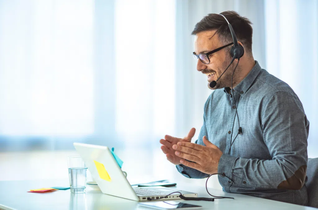 “Person wearing a headset speaking and gesturing while working on a laptop at a desk, with a glass of water and sticky notes nearby.”