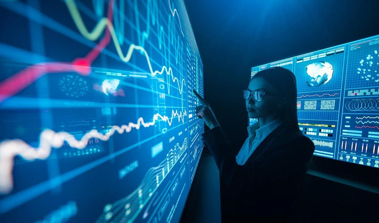 A person stands in a dark, high‑tech room, pointing at large blue digital dashboards filled with line charts, graphs, and world‑map data displayed across multiple wall‑sized screens.