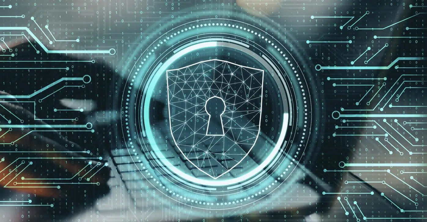 A futuristic cybersecurity-themed digital illustration showing a glowing shield with a keyhole icon at its center, symbolizing data protection. The shield is outlined with interconnected geometric lines, surrounded by circular HUD-style elements and binary code patterns. In the background, a blurred laptop keyboard is visible, while abstract circuit-like lines and dots extend across the image in shades of teal and blue, representing networks, encryption, and digital security systems.