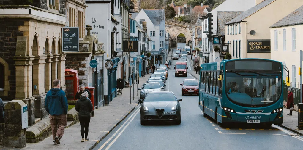 A sloping street in a small town lined with historic stone and pastel-colored buildings, with cars driving down the road and a teal bus in the foreground. Pedestrians walk along the pavement, passing shopfronts and a traditional red phone box, while a stone archway and trees are visible in the distance under a lightly clouded sky.