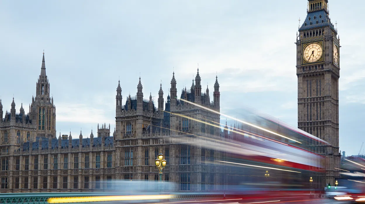 The Palace of Westminster and the Elizabeth Tower (Big Ben) in London at dusk, with a red double-decker bus passing across Westminster Bridge, creating motion blur against the historic Gothic architecture and cloudy sky.