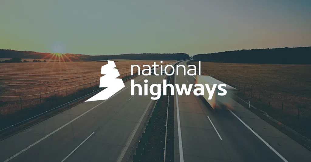 A wide, straight dual carriageway stretches through open countryside at sunset, with fields on either side and low hills in the distance. The sun is low on the horizon, casting warm orange light and long shadows across the landscape. Blurred vehicles travel along the road, suggesting motion. Overlaid in the centre is the white “national highways” logo and text, featuring a stylised map outline next to the words “national highways”.