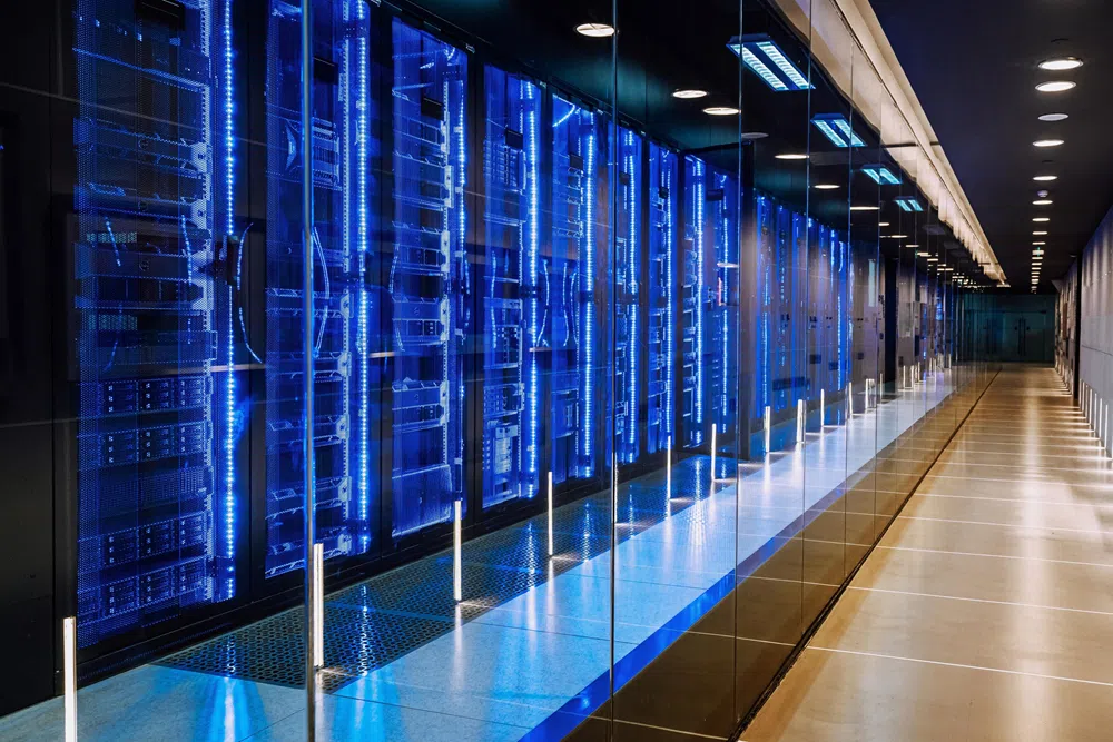 Modern data center server room with rows of glass-enclosed racks illuminated by blue LED lights, extending down a long, clean corridor.