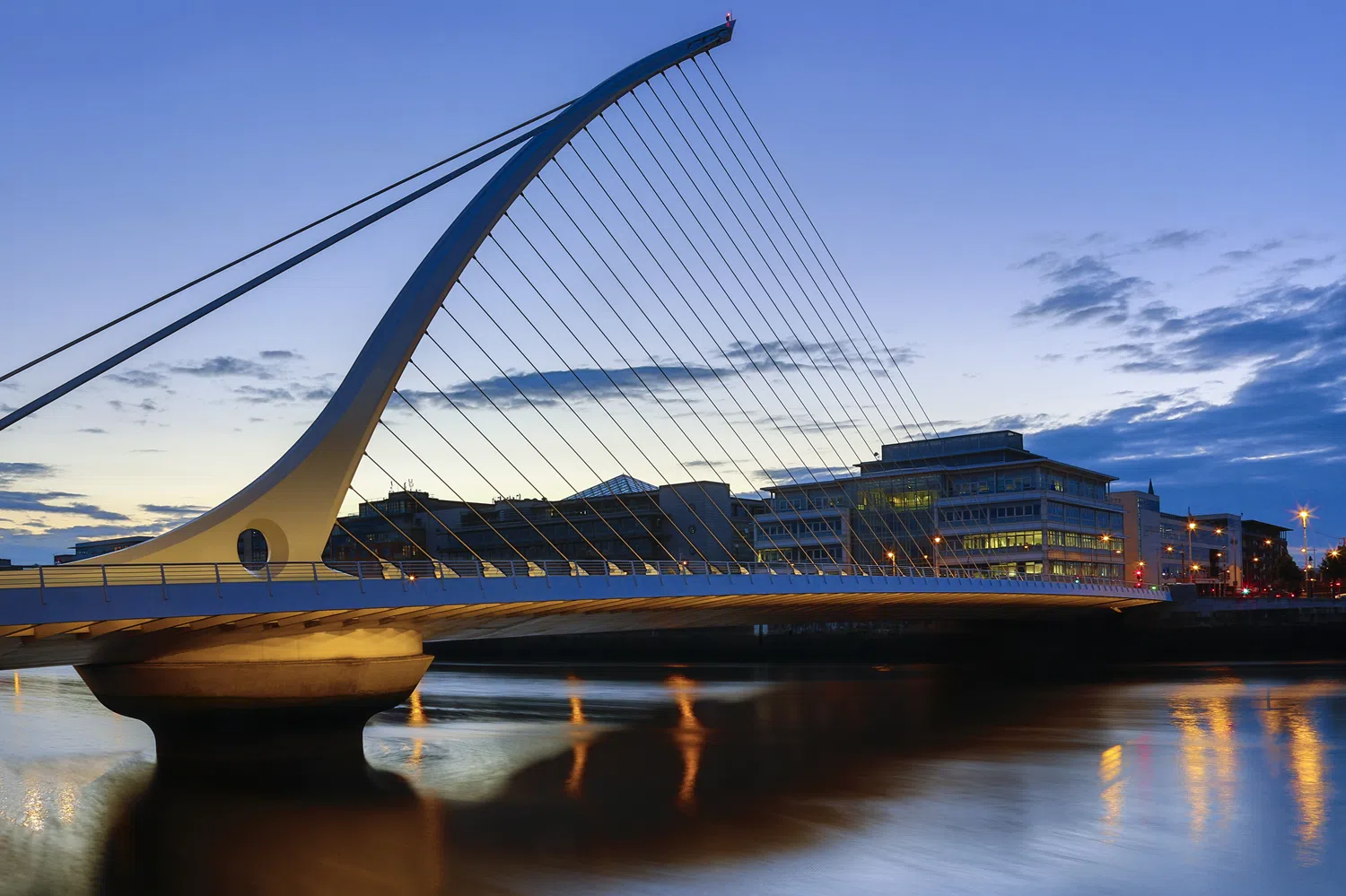 Samuel Beckett Bridge in Dublin, a modern cable-stayed bridge with a curved white harp-shaped pylon and cables spanning the River Liffey at dusk, with city lights reflecting on the water.