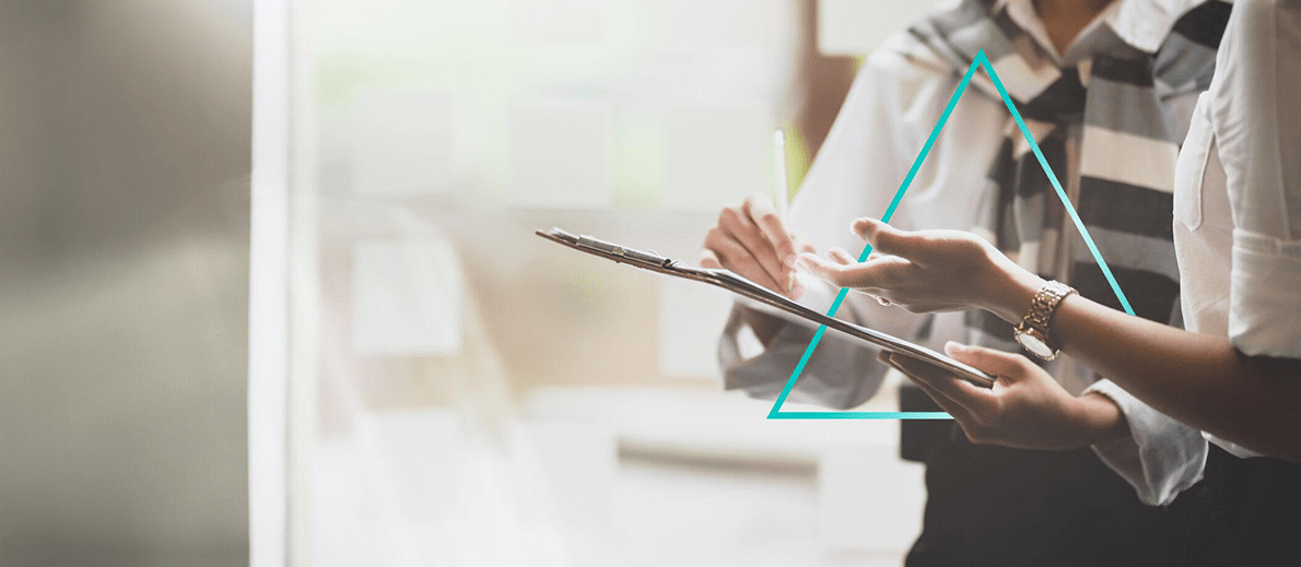 Two people collaborate while reviewing documents on a clipboard, one pointing with a pen as they discuss details. A geometric triangle graphic overlays the scene, adding a modern design element.