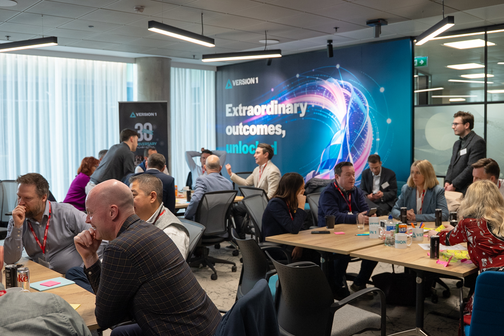 A busy workshop setting labeled “AI Co-Creation Studio Version 1”, showing multiple groups of people seated around tables collaborating with notebooks, sticky notes, and drinks. In the background, a large digital display reads “Extraordinary outcomes, unlocked” alongside a colorful abstract graphic. Additional signage includes “VERSION 1” and a separate poster with the number “38.” A teal triangular graphic overlay frames part of the scene, adding a modern, branded design element.