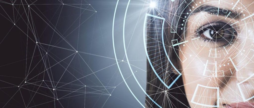 The image presents a close-up of part of a human face, focusing on one eye, overlaid with futuristic digital interface graphics. The eye is sharply in focus, while the rest of the face is partially cropped and softly blurred, drawing attention to the iris and surrounding area. Across the image, translucent geometric lines and nodes form a network-like pattern, suggesting data connections or artificial intelligence systems. Circular interface elements and targeting-style graphics are layered around the eye, resembling biometric scanning or facial recognition technology. The background transitions into a gradient of dark blue and grey tones, reinforcing a high-tech atmosphere. The combination of the human feature and digital overlays creates a visual theme centered on advanced technology, data analysis, and biometric identification systems.