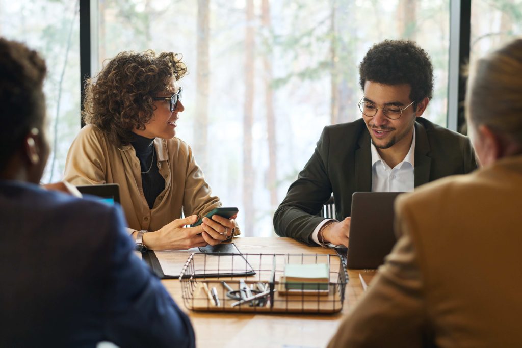 Four people sit around a table using a tablet and documents in a sunlit meeting room.