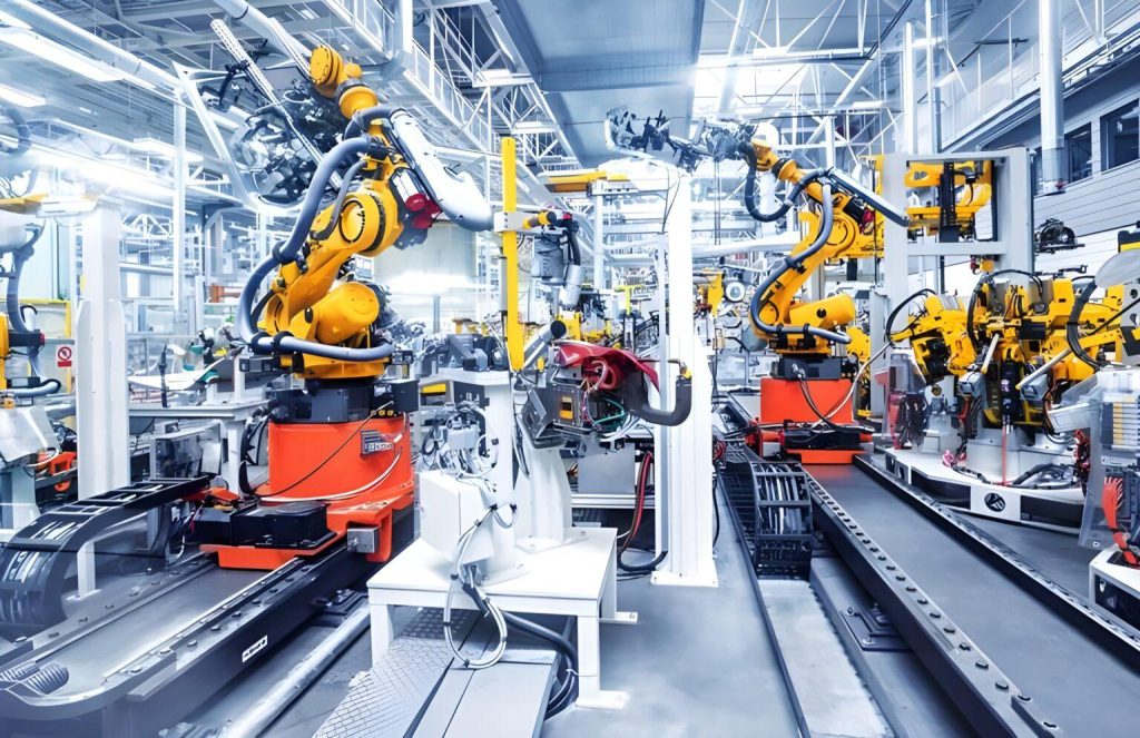 A high-tech industrial setting inside a modern factory, likely an automotive manufacturing facility. The scene is filled with multiple large robotic arms painted in bright yellow, each mounted on sturdy orange or red bases. These robotic arms have several joints and attached tools, allowing for precise, automated movement. In the foreground and center, one robotic arm is positioned near a partially assembled metal structure—possibly part of a vehicle frame—with visible wiring, tubing, and components attached. The robotic arm appears to be engaged in assembly or welding tasks. Additional robotic arms are arranged across the background on both the left and right sides, creating a dense, organized production line. Conveyor tracks and support rails run along the floor, indicating a system for moving parts through different stages of assembly. The factory interior is spacious and brightly lit with white lighting, supported by a network of metal beams and overhead structures. Numerous cables, pipes, and mechanical components are visible throughout, contributing to the complex and highly automated environment. The overall scene conveys advanced manufacturing processes, automation, and precision engineering, with no visible human workers present in the immediate frame.