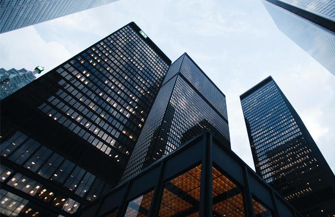 Low-angle view of several tall, modern office skyscrapers with dark, reflective glass façades. Three main buildings dominate the frame, rising sharply against a pale, overcast sky. The building on the left has a grid of illuminated windows scattered across its surface, while the central building reflects neighboring structures and the sky with a glossy finish. A third, slightly narrower tower stands to the right with evenly spaced window lights. In the foreground, part of a lower structure with a geometric, recessed ceiling pattern glows with warm orange lighting, contrasting with the cool blue and grey tones of the surrounding buildings.