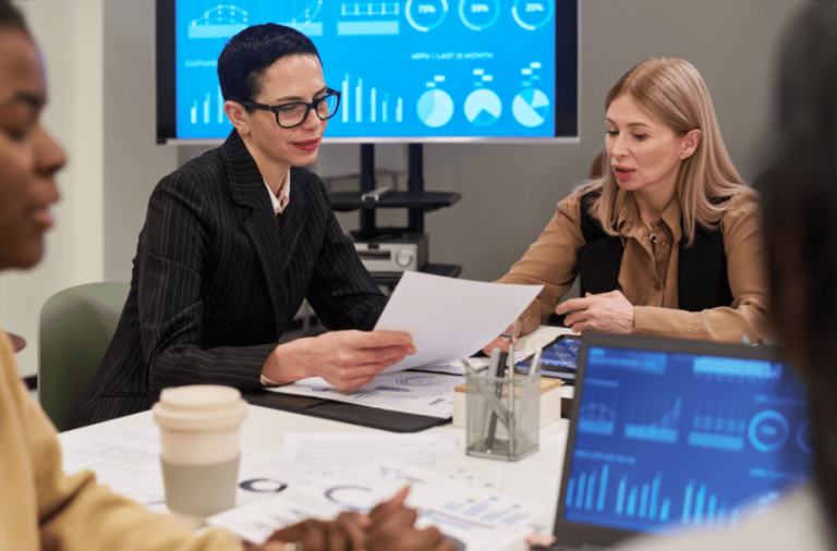 A group meeting in an office setting where several people sit around a table reviewing documents and digital data. In the center, one person in a dark striped blazer with glasses holds and reads a printed sheet, while another person beside them in a tan shirt and dark vest looks toward the paper and gestures with a hand. The table is covered with printed charts and reports, and a takeaway coffee cup sits near the foreground. A laptop displaying graphs and blue-toned analytics dashboards is partially visible at the lower right. In the background, a large monitor shows multiple blue charts, including bar graphs, line graphs, and pie charts. The lighting is bright and even, with neutral-colored walls contributing to a professional office environment.