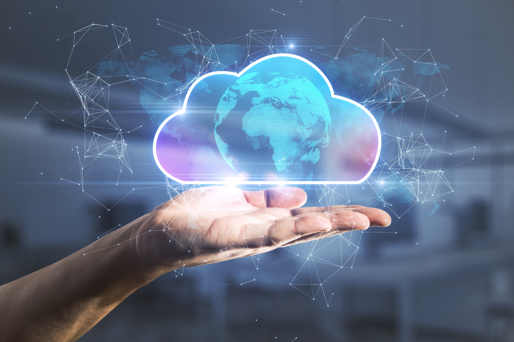 A close-up of a human hand held open with a glowing cloud icon hovering above the palm. Inside the cloud shape is a semi-transparent globe showing continents, symbolising global cloud computing. The background features a blurred office-like setting with a network of light lines and nodes, representing digital connectivity and data flow. The overall colour palette includes cool tones of blue and purple with bright highlights around the cloud.