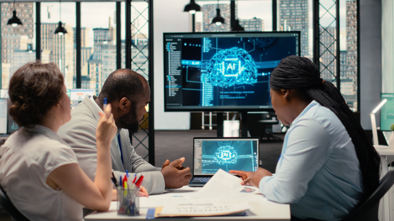 Three people sit around a table in a modern office with large windows and a city skyline visible in the background. In the center of the table, a laptop displays a visual interface featuring an abstract brain graphic labeled “AI,” surrounded by data elements. Behind the group, a large monitor shows a similar artificial intelligence-themed interface with code panels and a glowing brain visualization. Papers and pens are spread across the table, and the individuals appear to be discussing or reviewing the information on the screens, suggesting a collaborative meeting focused on AI or data analysis.Provide your feedback on BizChat