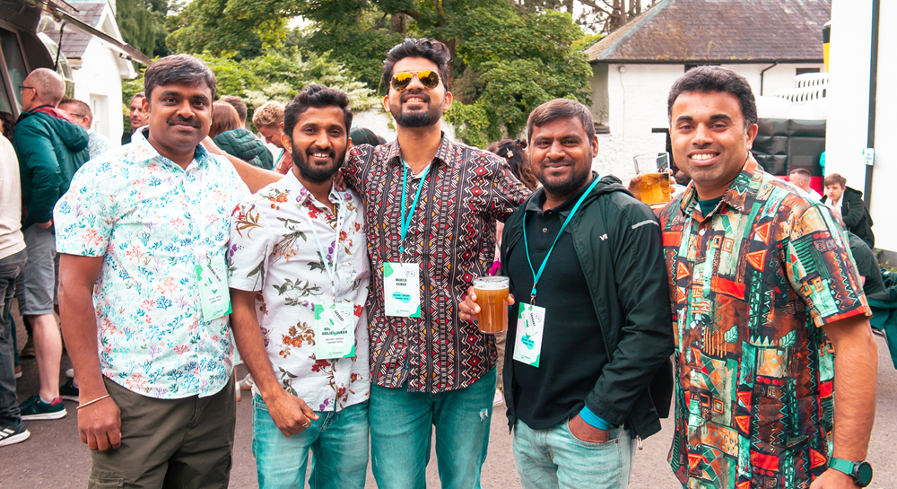A group of five adults stand together outdoors at a casual networking or social event, posing for a photo. Each individual is wearing a lanyard with a visible event badge, suggesting a professional or careers-related gathering. Several of the individuals are dressed in patterned short-sleeve shirts, creating a relaxed and informal atmosphere. One individual near the center is holding a plastic cup filled with a drink. The group stands closely together in the foreground, facing the camera. Behind the group, more attendees are visible in a busy