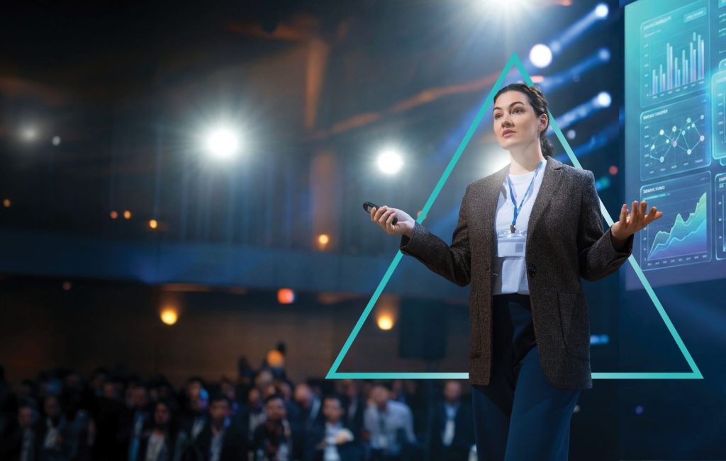 A conference speaker stands on a stage holding a presentation remote, addressing an audience. Behind the speaker, a large screen displays multiple SaaS-style data dashboards with line graphs and charts, while event lighting and a geometric overlay triangle graphic frame the scene.