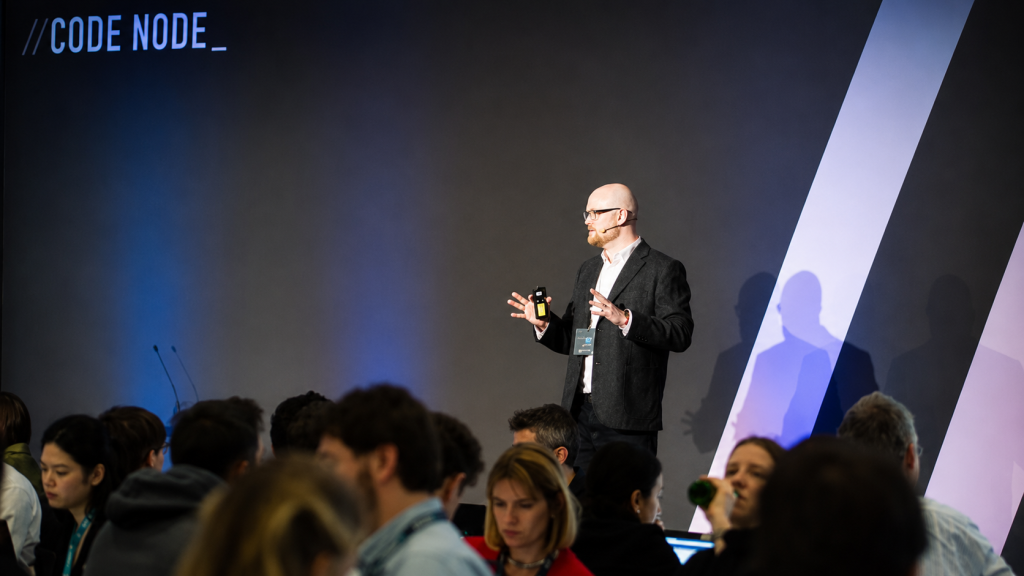 Conference speaker presenting on a stage at a technology event. The presenter stands in front of a dark backdrop with large diagonal light-colored design elements, holding a remote device while addressing an audience seated in the foreground. The audience fills the lower portion of the image, with some attendees using laptops. Blue stage lighting illuminates the scene, and the text “//CODE NODE_” appears in the upper-left corner.
