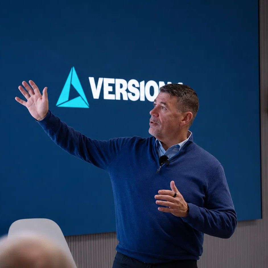 Man presenting in a modern conference room, gesturing toward a large screen displaying financial services icons and text, with a corporate logo on a blue background behind him
