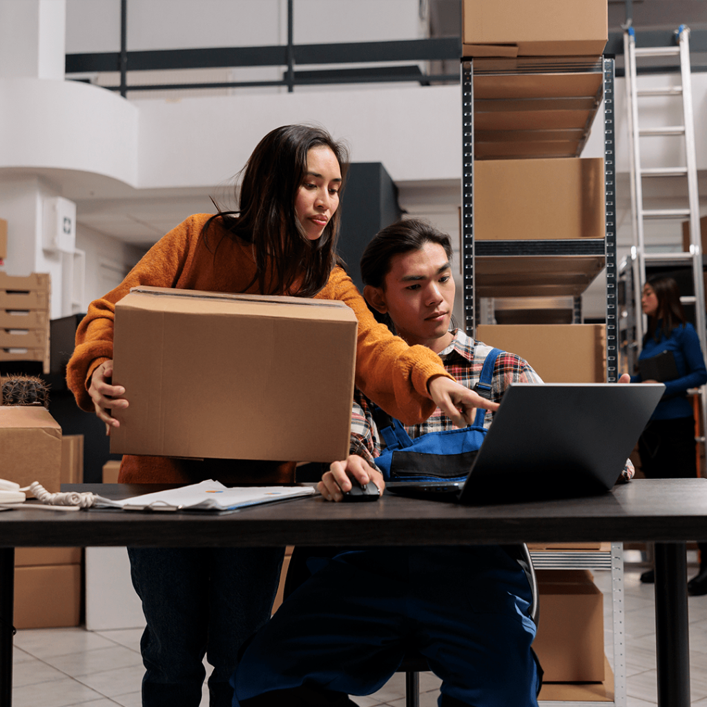 Two people, looking at a laptop, in a warehouse, one carrying a box