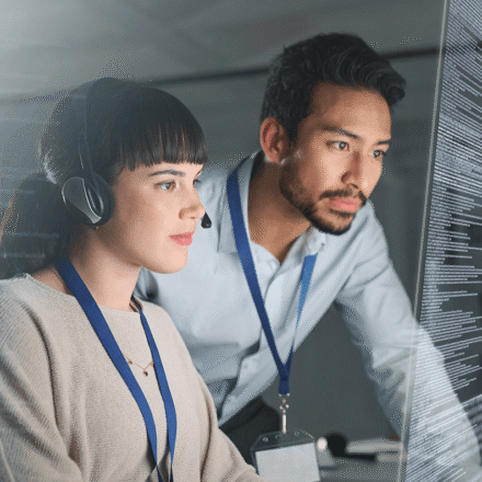 Two professionals wearing lanyards work closely together at a computer workstation. One person is seated and wearing a headset while looking at a large monitor displaying lines of code. A second person leans in from the side, also focused on the screen. The setting appears to be a modern office or tech environment with soft lighting, and the image conveys collaboration around software development or data analysis.