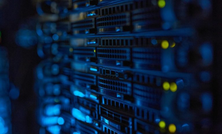 Close-up view of stacked server racks in a data center, illuminated by blue lighting with small green and yellow status lights visible on the hardware.