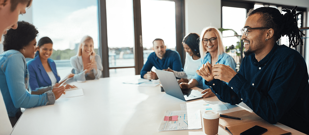 A group of people sit around a conference table in a brightly lit meeting room, discussing ideas with laptops, documents, and coffee cups in front of them.