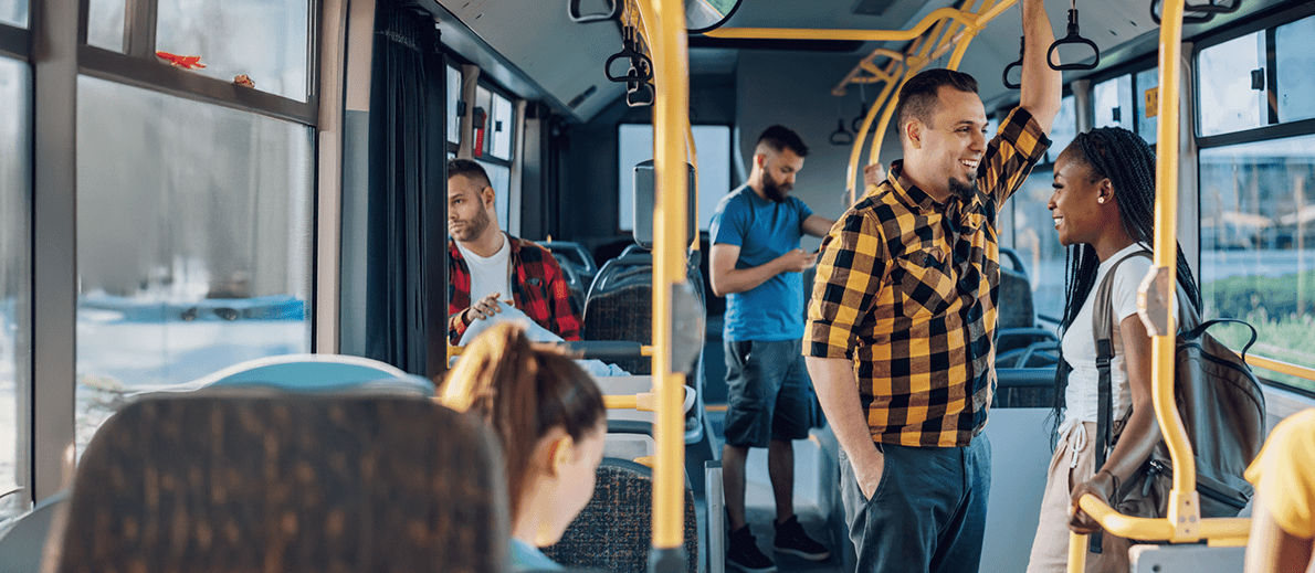 A photograph taken inside a public bus during daylight, showing several passengers and the interior layout. In the foreground, there are yellow vertical and horizontal handrails typical of public transport seating areas. Near the center-right, one passenger stands facing another passenger. The standing passenger is holding onto an overhead rail with one hand while wearing a yellow-and-black checkered long-sleeve shirt and dark trousers. The other nearby passenger, standing and facing toward the first, has long braided hair and is wearing a light-colored top with a backpack over one shoulder. In the background, additional passengers are seated and standing. On the left side, one seated passenger is wearing a red plaid shirt and looking downward at an object held in both hands. Further back in the aisle, another standing passenger is wearing a blue T-shirt and also looking down at a handheld device. The bus interior includes rows of grey seats, overhead hand grips, and large windows along the side allowing natural light into the space. Through the windows, an urban outdoor setting is faintly visible but not clearly detailed. The scene shows a typical moment of public transit use, with passengers standing, sitting, and holding onto support bars while the bus is in motion.
