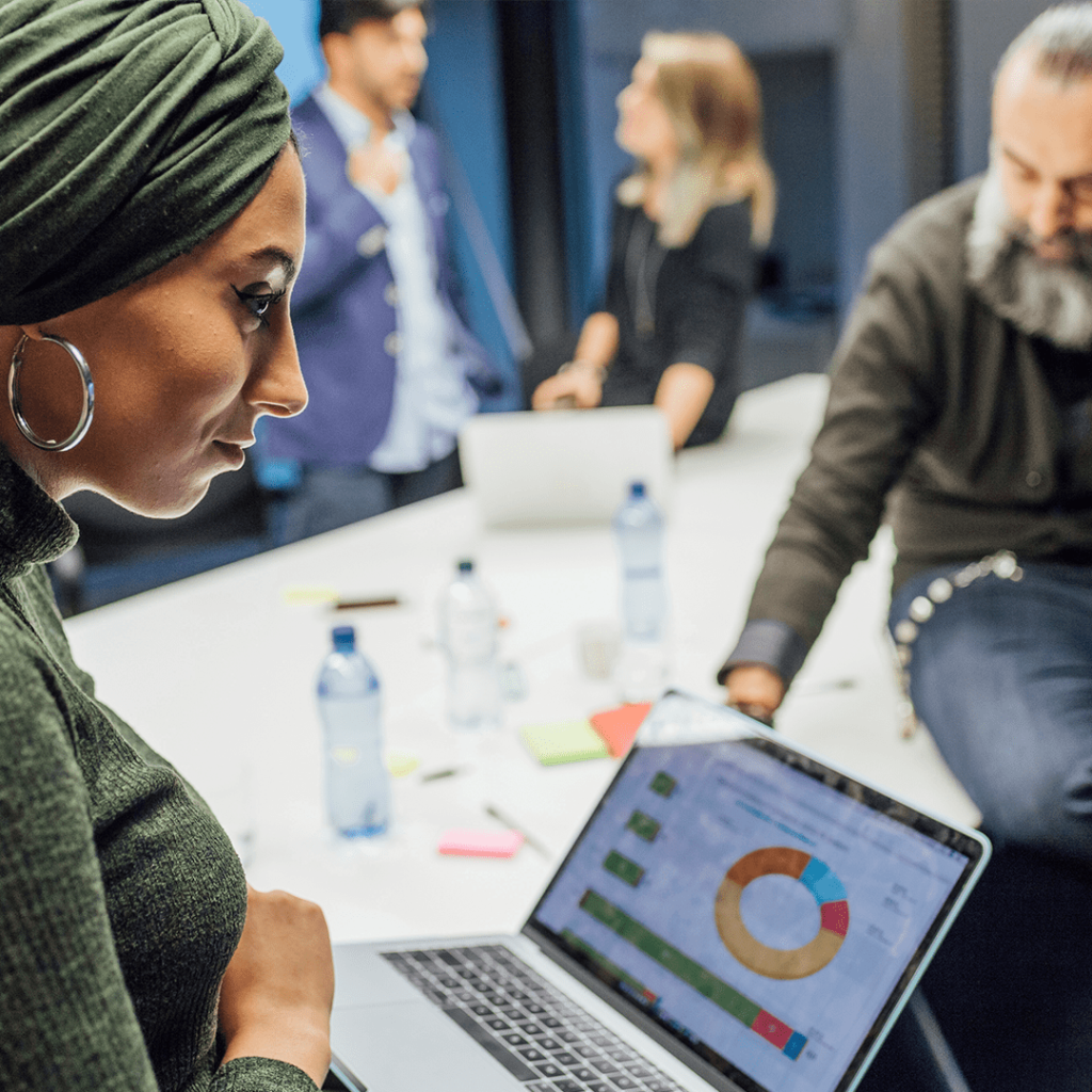“Woman wearing a headscarf reviews analytics on a laptop during a team meeting, with colleagues discussing in the background.