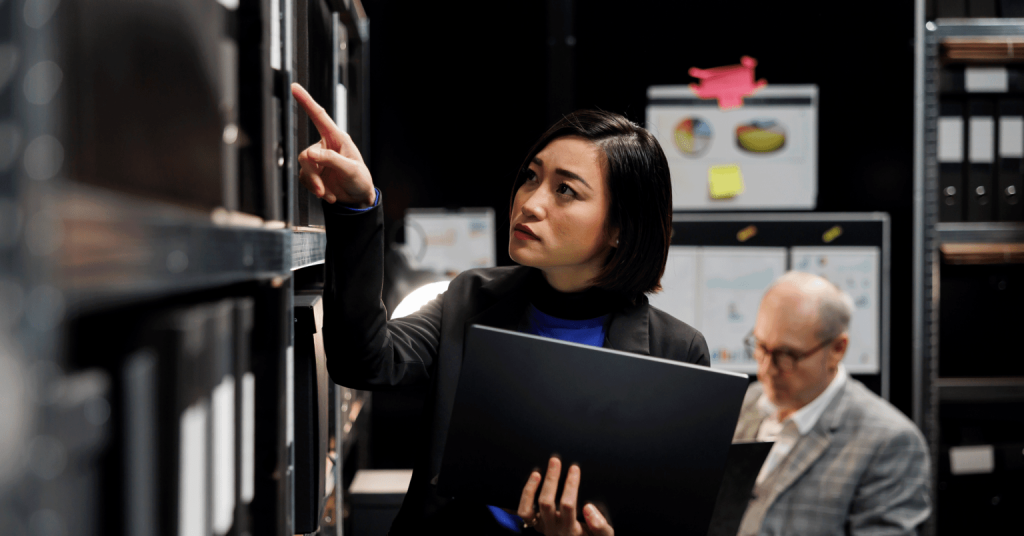 A close-up indoor scene showing a person interacting with equipment in what appears to be a technical or office environment. The person is holding a closed or partially open laptop in the left hand while reaching upward with the other hand to point at a vertical surface or panel mounted on a rack or cabinet. The person is dressed in a dark blazer over a blue top, suggesting a professional setting. The individual’s hand is extended with the index finger pointing toward a specific spot on the vertical panel, indicating focus on a particular control, screen, or component. In the foreground on the left side, there is a dark vertical structure resembling a server rack, equipment cabinet, or shelving unit with electronic or mechanical components inside. The person is positioned very close to this structure, interacting with it directly. The background is softly blurred but includes a whiteboard or display area with charts or diagrams, as well as a few brightly colored sticky notes attached to a surface. These elements suggest a workspace used for planning, monitoring, or analysis. The lighting is warm and even, highlighting the person and the equipment clearly while keeping the background less prominent. The overall scene conveys a situation involving inspection, troubleshooting, or management of technical systems or data. Provide your feedback on BizChat