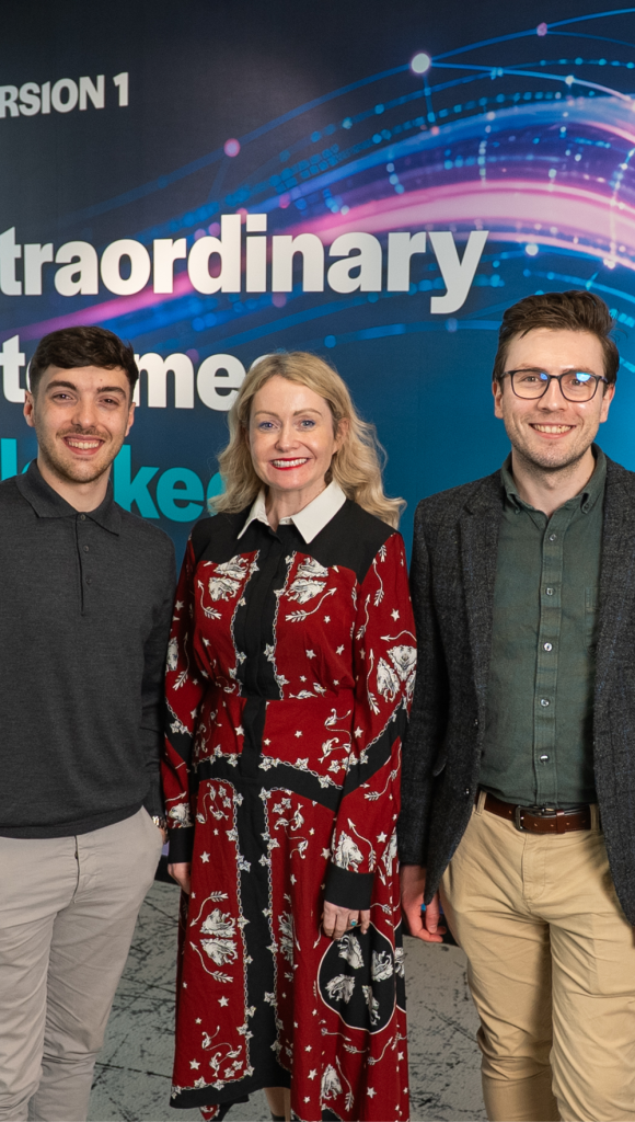 Three professionally dressed adults stand side by side, smiling at the camera in front of a blue event backdrop with abstract digital wave graphics and partial text.