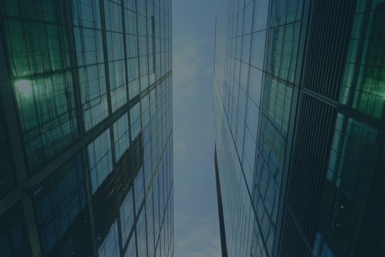 View looking upward between two tall, modern glass office buildings that rise on the left and right sides of the frame. The buildings have reflective glass façades with a grid of window panels, tinted blue-green, and they mirror parts of the surrounding city. The perspective creates strong vertical lines that converge toward a narrow strip of pale sky in the center. The overall scene feels symmetrical and geometric, with clean lines, cool tones, and a slightly overcast sky visible above.