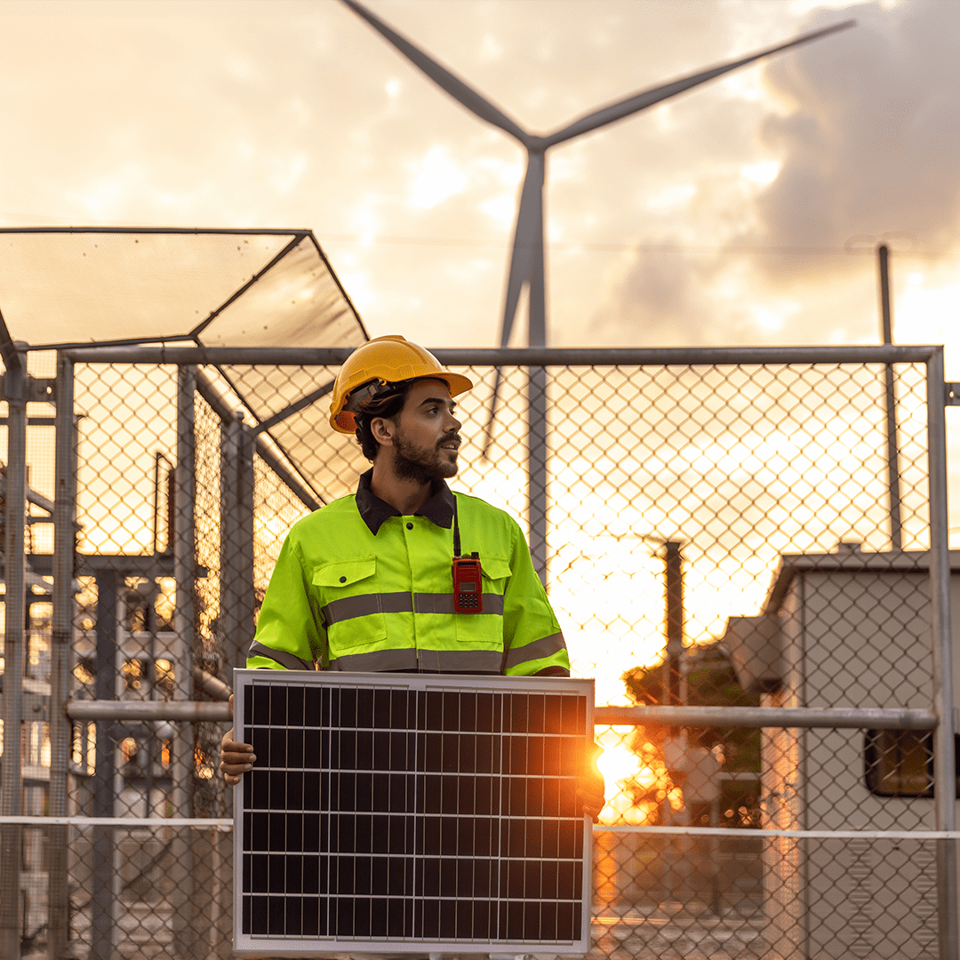 A photograph taken at a renewable energy site during sunset. In the foreground, a construction or maintenance worker stands facing slightly to the left, holding a rectangular solar panel upright with both hands. The worker is wearing a bright high-visibility yellow jacket with reflective strips and a yellow hard hat. A small device, likely a radio or communication unit, is clipped to the front of the jacket. Behind the worker, a metal fence encloses industrial equipment and structures, suggesting a secured energy or utility facility. In the background, a large wind turbine rises into the sky, with its blades extending outward and partially silhouetted against the light. The sky is filled with warm, golden tones from the low sun, which is positioned near the horizon and shines through the fencing, creating a bright flare near the bottom right of the solar panel. The combination of the solar panel and wind turbine highlights renewable energy technologies such as solar and wind power.
