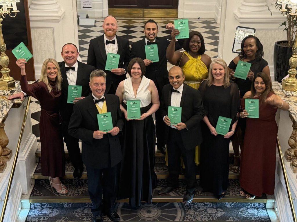 A formal group photo of around eleven adults standing on the steps inside an elegant building with a black-and-white checkered floor and white architectural columns. Each person is dressed in formal evening attire such as suits, tuxedos, and long dresses. All individuals are holding up or displaying green booklets or certificates featuring a white circular logo and small white text on the cover. The group is arranged in two rows, with some people standing slightly higher on the steps behind others. The setting appears upscale, with polished stone flooring, decorative railings, and a symmetrical layout leading to a doorway in the background. The lighting is bright and even, highlighting the group and their certificates. The overall composition suggests a celebratory or award-related event where participants are being recognized or honored.