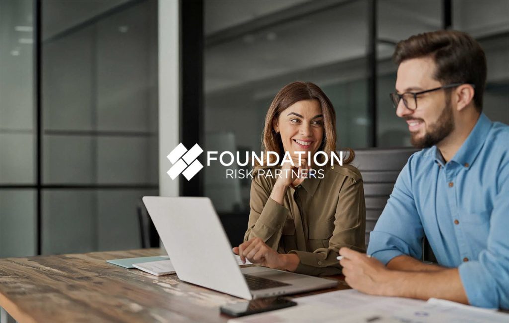 Two colleagues standing in a modern office discuss something on a tablet. A woman wearing a beige hijab and light blazer looks at the screen while a man in a light blue shirt holds the tablet and speaks. The overlay reads “Foundation Risk Partners” with the company logo.
