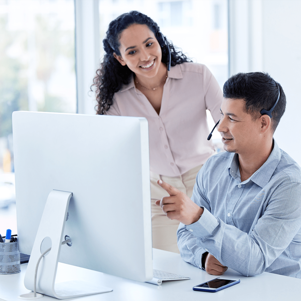 Two office workers wearing headsets collaborate at a desk, looking at a large desktop monitor, with one pointing at the screen in a bright, modern workspace.