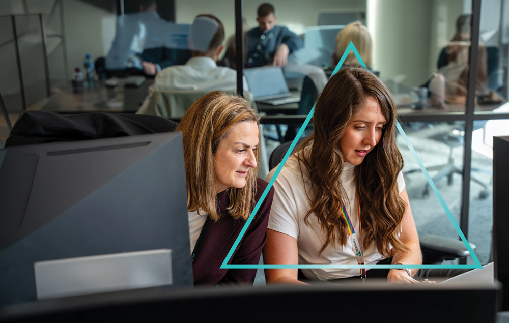Two women sit closely together at a workstation, focused on a computer screen as they collaborate, with a modern office and glass meeting rooms visible in the background, and a subtle triangular graphic overlay framing them.