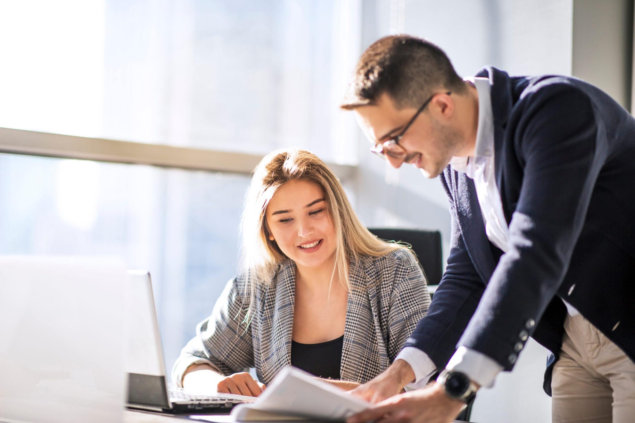 Two people in a bright office collaborate at a desk, reviewing documents while one points and explains.