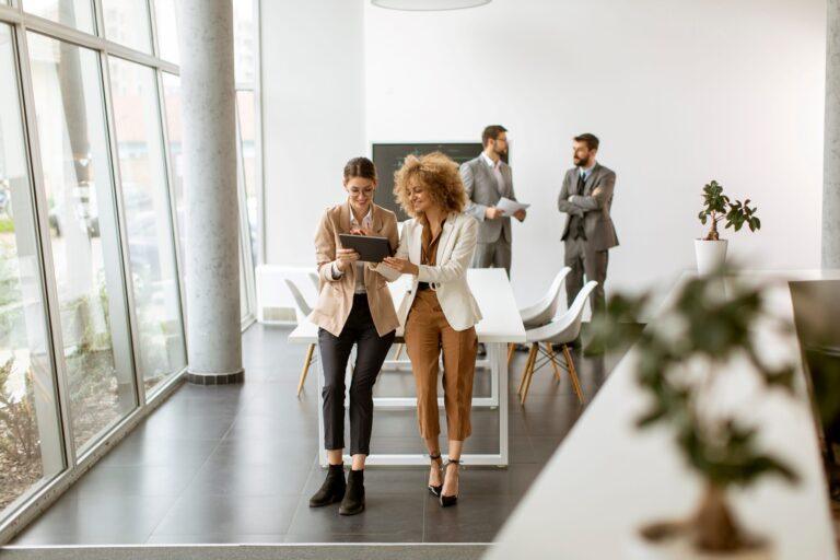 The image shows a modern office interior with a clean, minimalist design and large floor-to-ceiling windows allowing abundant natural light. In the foreground, two individuals walk side by side across an open space while closely examining a tablet device that one of the individuals holds. Both individuals are dressed in smart business attire, and their posture suggests an active discussion or review of information on the screen. Behind them, a large white table with several chairs is positioned centrally, indicating a meeting or collaboration area. Further in the background, two additional individuals stand near the table, facing each other and appearing engaged in conversation while holding documents. The space features light-colored floors, white furniture, and simple décor, including a small plant visible to the side, contributing to a bright, professional, and contemporary work environment.