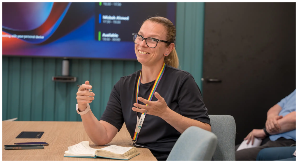 A staff member sits at a meeting table in a modern office, holding a pen and gesturing while speaking. A notebook and smartphone lie on the table in front of them. The staff member wears glasses, a dark top, and a rainbow lanyard with an ID badge. Behind the staff member, a large screen displays blurred presentation content, and another seated person is partially visible to the side.