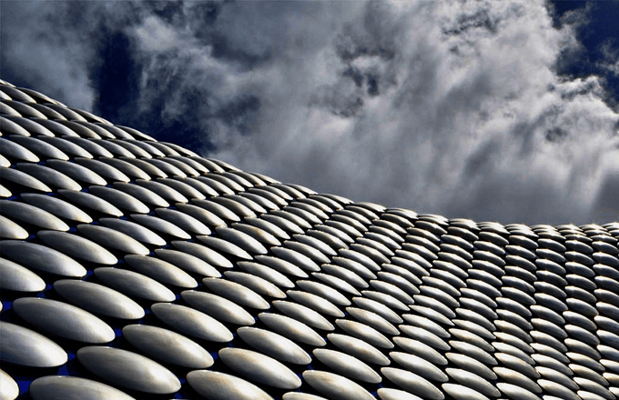 Close-up, low-angle view of a modern building exterior covered in thousands of smooth, oval, metallic-silver panels arranged in a repeating, scale-like pattern. The curved façade sweeps diagonally across the frame from lower right to upper left, emphasizing a wavy, organic architectural form. Above the structure, a dramatic sky with thick, textured clouds fills the background, creating contrast between the reflective, geometric surface and the soft, natural cloud formations.
