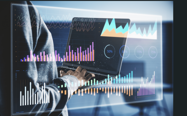 A close-up view of a person working on a laptop computer, overlaid with multiple transparent data visualizations. The laptop screen displays colorful charts, including vertical bar graphs in pink, purple, and orange, as well as a line graph and a mountain-style area chart in teal and light blue. A circular dashboard element shows “50%” in the center. Additional futuristic interface elements and waveform-like graphics appear layered across the image. The scene is lit with cool blue tones, giving a high-tech, analytical atmosphere. The person’s hands are positioned on the keyboard, suggesting active data analysis or monitoring.
