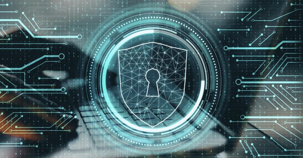 A futuristic cybersecurity-themed digital illustration showing a glowing shield with a keyhole icon at its center, symbolizing data protection. The shield is outlined with interconnected geometric lines, surrounded by circular HUD-style elements and binary code patterns. In the background, a blurred laptop keyboard is visible, while abstract circuit-like lines and dots extend across the image in shades of teal and blue, representing networks, encryption, and digital security systems.